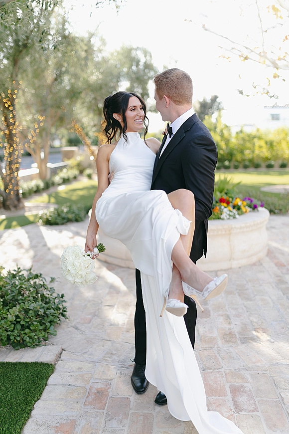 Couple portrait of groom carrying the bride as they laugh, her white gown and rose bouquet against garden greenery and string lights