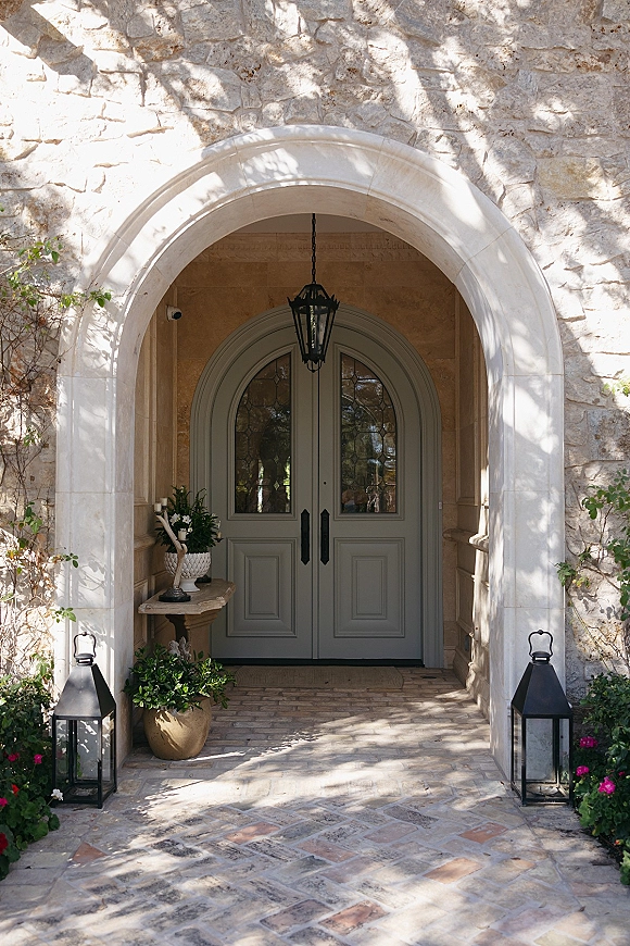 Wedding venue entrance with arched stone doorway and double door entryway, lit by hanging and floor lanterns beside candles and greenery
