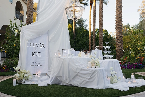 Wedding welcome sign with white fabric draping, crystal chandelier, candles and white florals beside a champagne tower in an outdoor garden setting