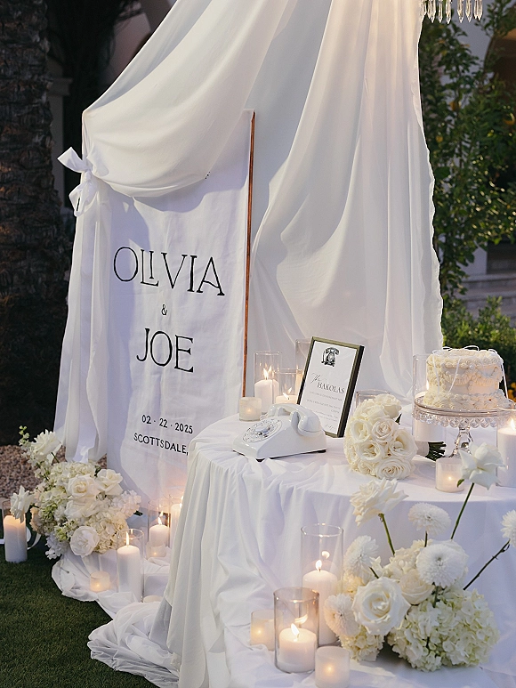 Wedding welcome table with fabric banner and framed sign, white rotary phone, candles, and white roses beside a small cake on a lawn backdrop