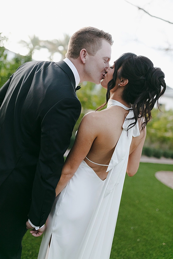 Wedding kiss portrait of bride and groom kissing, groom in black tuxedo, bride in backless halter gown with ribbon tie on sunlit lawn