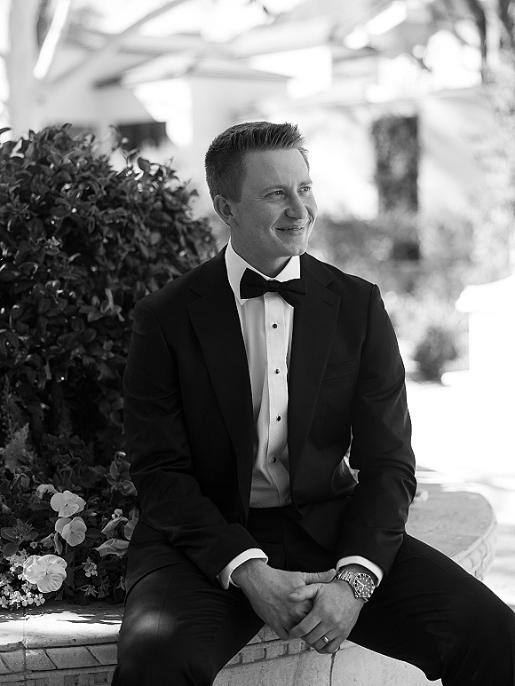 Groom portrait in tuxedo with bow tie and wristwatch, seated on a stone bench in a garden courtyard, smiling and looking away