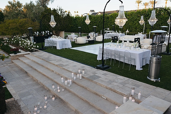 Outdoor reception decor with long banquet tables, clear ghost chairs, crystal chandeliers, and candlelit lounge seating on a garden lawn