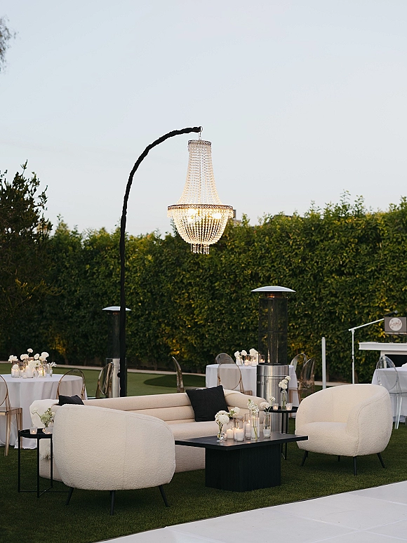 Wedding lounge decor with an outdoor chandelier over white sofas, candles and bud vases, set on a lawn patio beside a hedge wall.