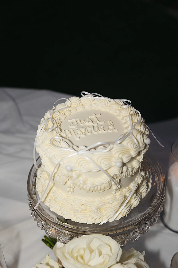 Wedding cake with white buttercream wedding cake piping, rosettes and sugar pearls, topped with ribbon bows on a stand against a dark backdrop