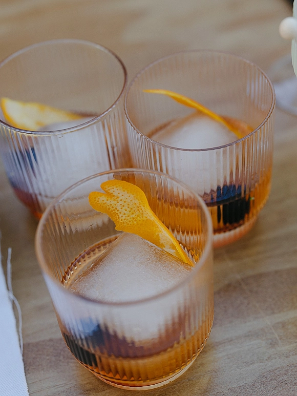 Wedding cocktails in rocks glasses with amber old fashioned, large ice cubes, and orange peel garnish on a wood tabletop