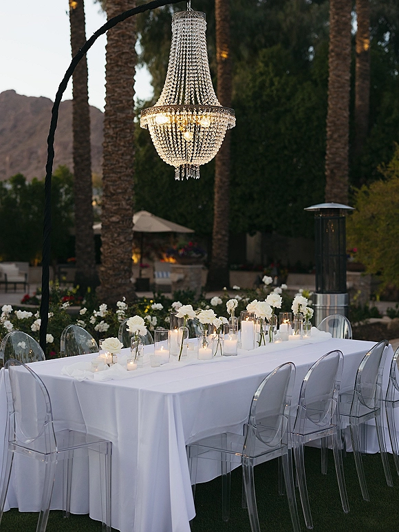 Reception tablescape with outdoor reception table under a crystal chandelier, white linens, taper candles, white flowers, and palm-lined patio views