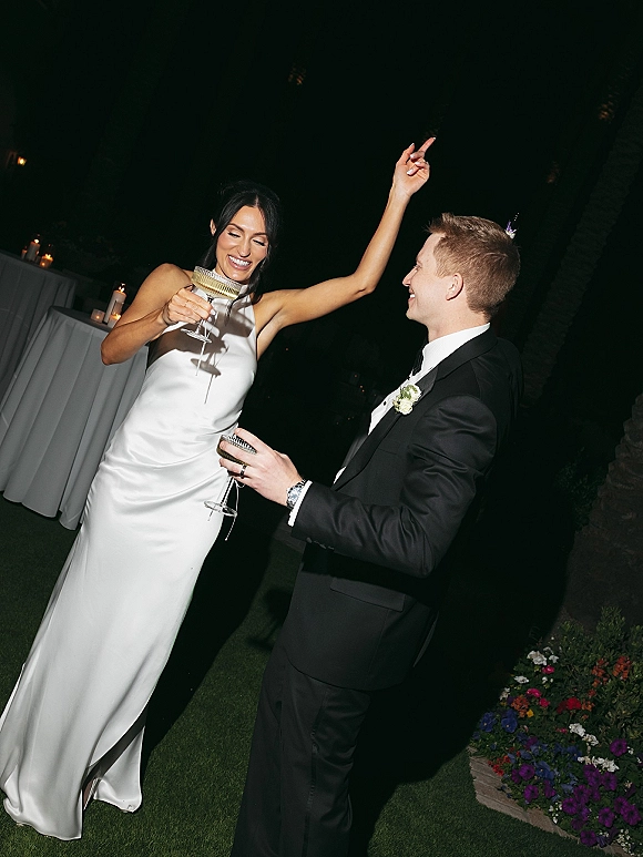 Wedding reception moment as newlyweds raise champagne coupe glasses, bride in satin dress and groom in tux at candlelit outdoor tables under night sky
