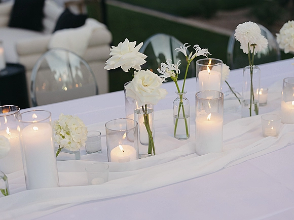Reception tablescape with wedding table centerpiece of white roses, hydrangea and floating candles on a gauze runner, acrylic chairs behind