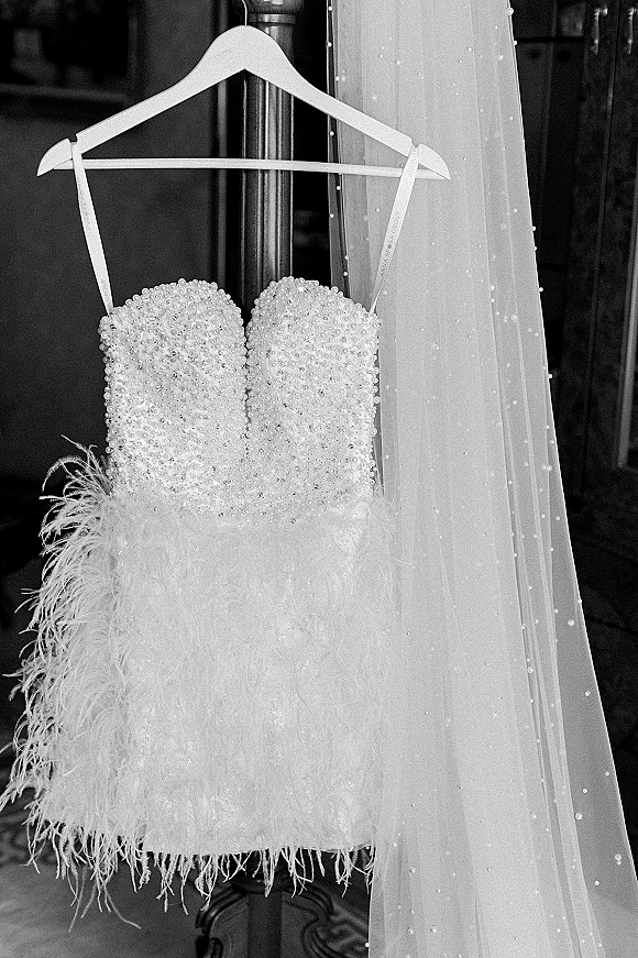 Wedding dress with pearl beading, a strapless silhouette and feather skirt hangs on a hanger in an indoor doorway on a wooden floor