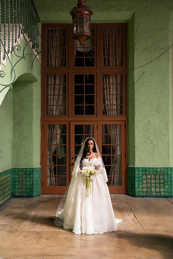 Bridal portrait of a bride in an off the shoulder wedding dress with veil and calla lily bouquet, standing in an arched doorway with lantern backdrop