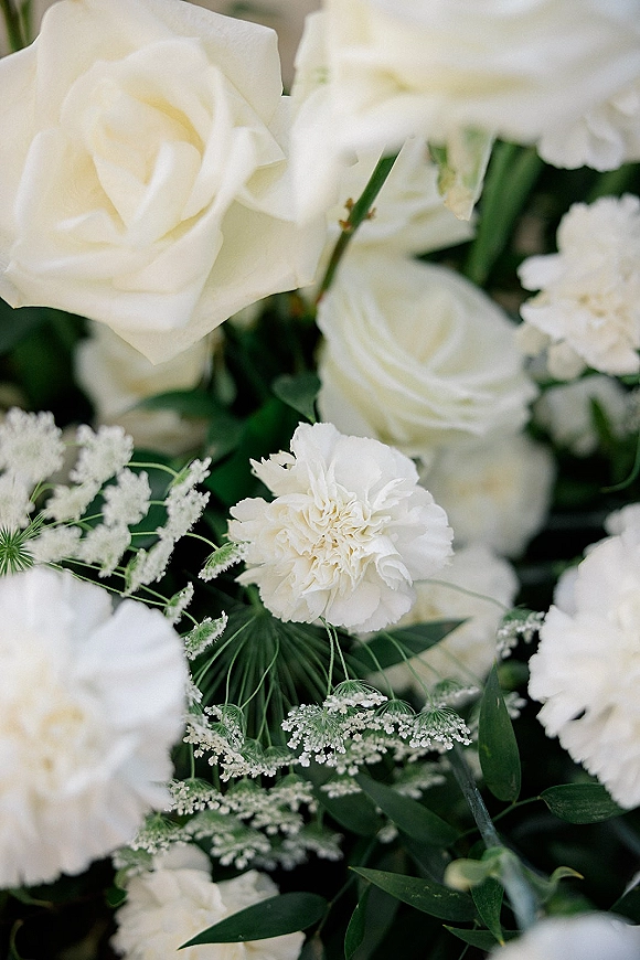 White wedding florals featuring white rose bouquet blooms with greenery foliage, clustered close-up against softly blurred florals and dark leaves