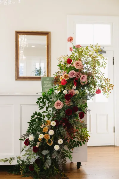 Wedding bar florals with a lush greenery and rose arrangement spilling over the bar counter, framed by a mirror and chandelier in a classic white room