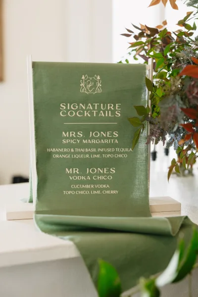 Wedding bar sign with signature cocktails sign on green fabric in an acrylic holder, framed by floral arrangement on a bar counter