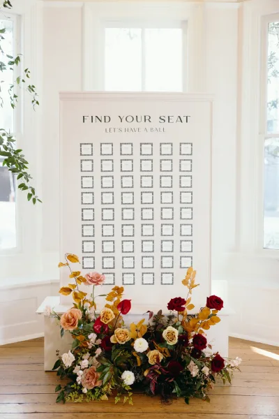 Wedding seating chart with find your seat sign and escort cards on a white plinth, accented by roses and greenery in a bright windowed room
