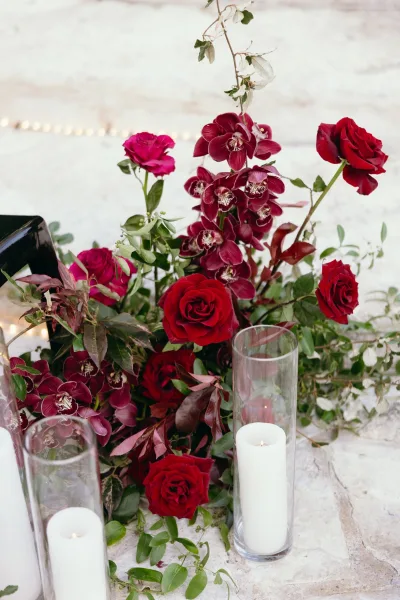 Wedding floral arrangement with red rose centerpiece and burgundy orchids, eucalyptus greenery, and pillar candles in glass vases against white brick wall