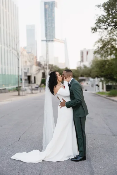 Wedding kiss portrait of bride and groom kissing on a city street, her long veil and dress train flowing as he holds her waist