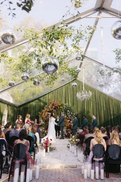Wedding ceremony under a clear top tent with couple at altar beneath floral arch, candle-lined aisle, greenery garlands, and chandelier accents