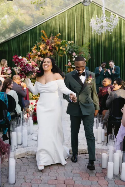 Wedding recessional as newlyweds walking up aisle, bride holds bouquet high past glass cylinder candles in an outdoor garden ceremony