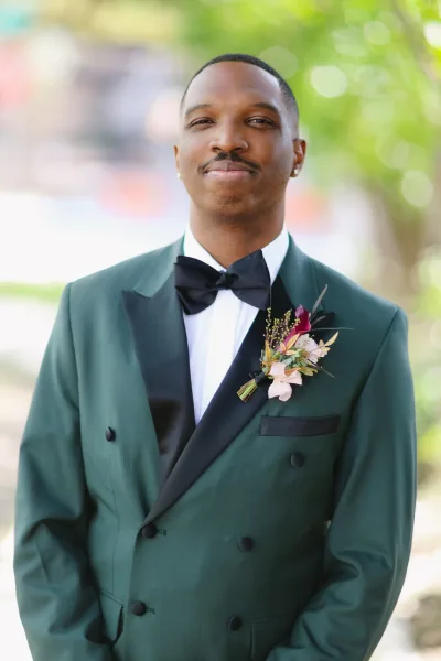 Groom portrait in an emerald green tuxedo with black bow tie and floral boutonniere, standing against blurred outdoor greenery in daylight