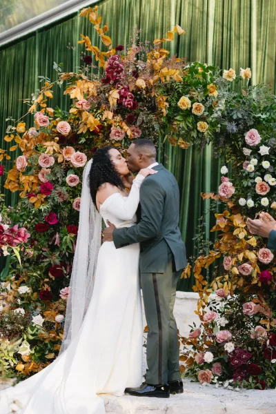 Wedding kiss as bride in off the shoulder dress and long veil kisses groom in green suit beneath a rose and greenery arch on stone steps