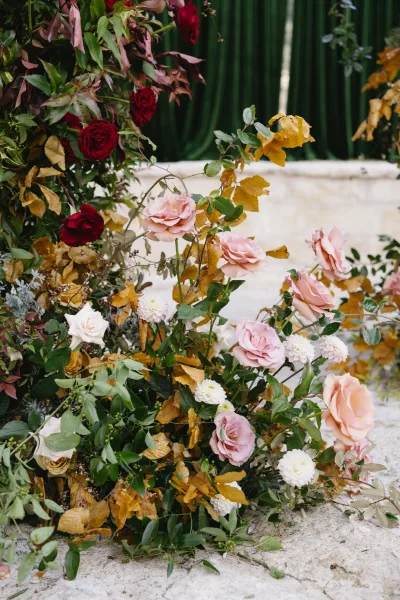 Wedding floral arrangement with roses and dahlias spilling with greenery against a white stone wall and green drapery backdrop
