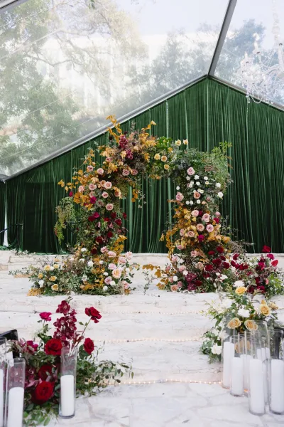 Wedding ceremony backdrop with an asymmetrical floral ceremony arch of roses and greenery, velvet drape, candles, chandelier, and string lights in a clear tent