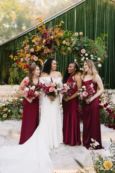 Bride with bridesmaids in burgundy holding bouquets beneath an asymmetrical floral arch with greenery and string lights in a garden setting