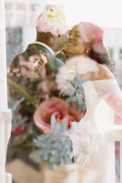 Wedding couple portrait of bride and groom embrace, groom kissing her cheek as she smiles in window light with floral blur foreground