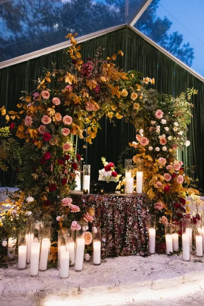 Wedding sweetheart table with a rose floral arch, sequin tablecloth, and pillar candles before a green velvet drape under a glass tent ceiling