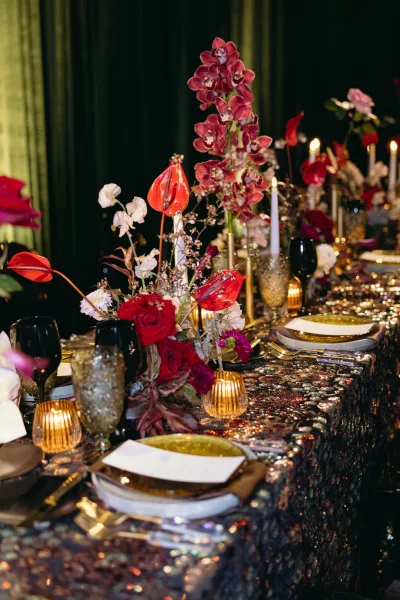 Reception tablescape with sequin wedding tablecloth, red orchids and roses, gold candleholders, taper candles, and smoked goblets against dark green drapery