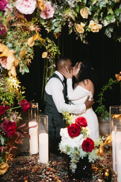 Wedding kiss portrait of bride and groom kissing beneath a floral arch with greenery and roses, framed by pillar candles against a black drape backdrop