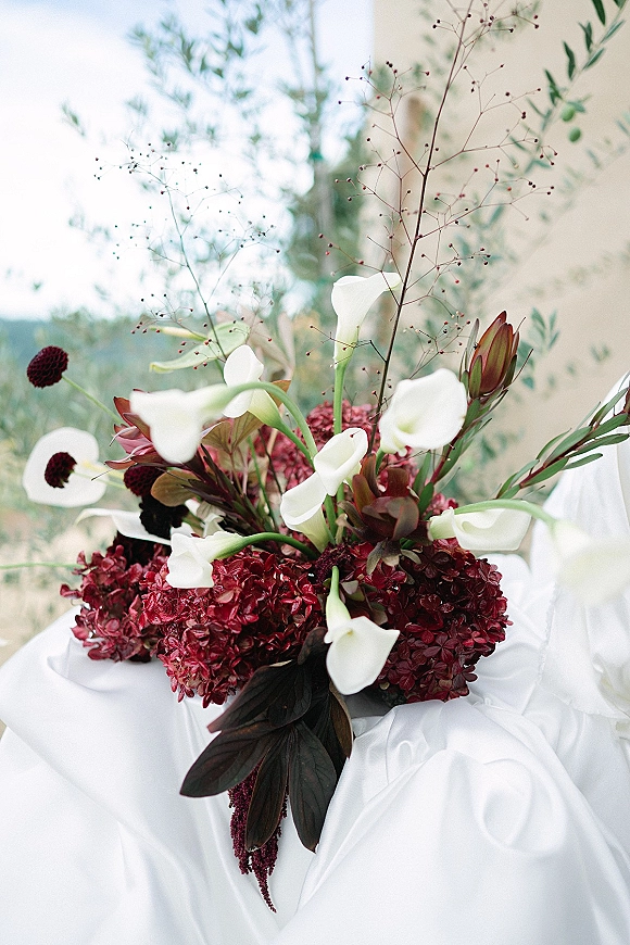 Wedding bouquet with calla lily bouquet blooms, burgundy hydrangea and protea, framed by dark foliage against soft greenery and mural backdrop