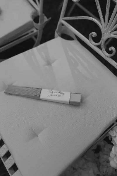Wedding favor folding fan with a personalized label resting on a metal chair cushion, set among flowers in an outdoor ceremony space