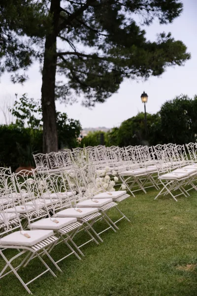 Ceremony seating with outdoor wedding ceremony chairs in rows of white wrought iron, cushioned seats, programs, and aisle florals on a grassy lawn under a large tree