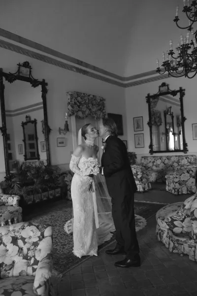 Wedding kiss portrait of bride and groom kissing, her long veil and bouquet beside a floral sofa under chandeliers in an indoor lounge