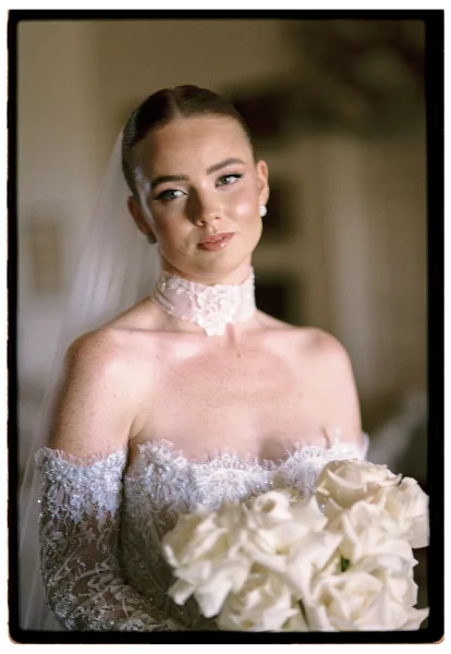 Bridal portrait of a bride holding bouquet of white roses, wearing an off-the-shoulder lace dress and veil against a neutral indoor wall