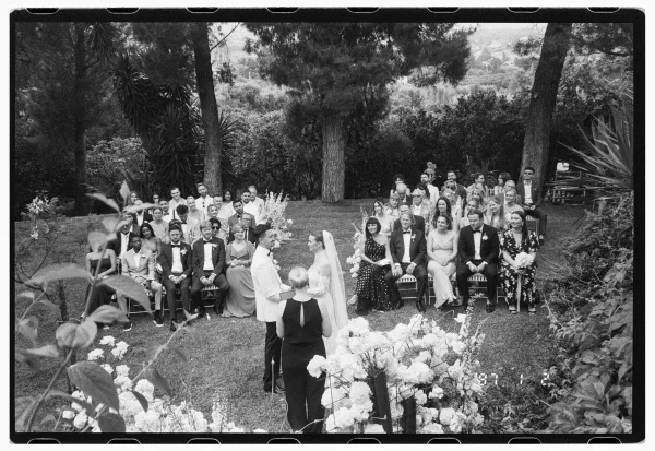 Ceremony moment as bride in a long veil and groom in a light suit face the officiant on a garden lawn with seated guests and flowers