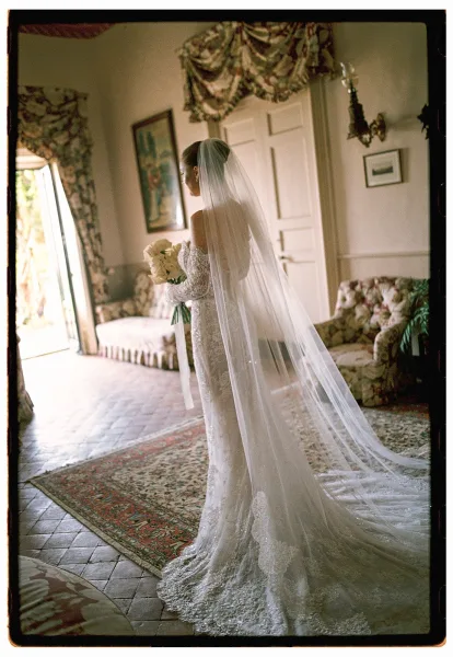 Bridal portrait of a long veil bride holding a white bouquet with ribbon wrap in a vintage sitting room with floral curtains and sofa