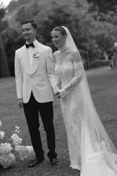 Wedding couple portrait in black and white, bride laughing in a lace high-neck gown and long veil beside groom in white jacket on garden lawn