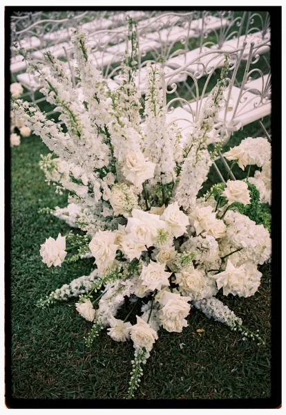 Ceremony aisle flowers in lush white roses, hydrangea, and delphinium with greenery beside white wrought iron chairs on a grass lawn