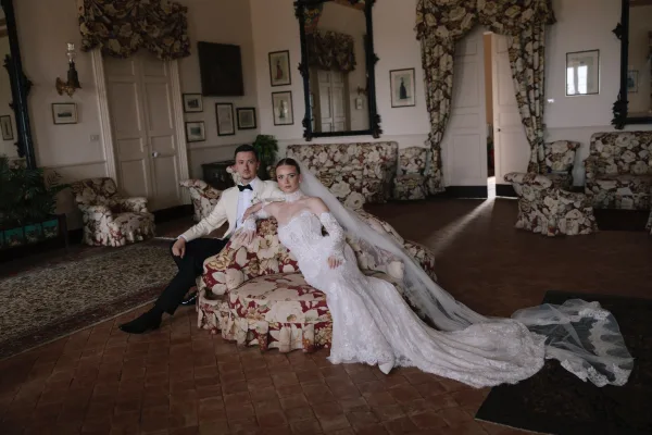 Couple portrait of bride and groom sitting on a floral sofa, her cathedral veil pooling on tiled floor in a vintage sitting room