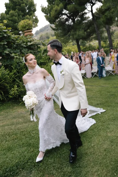 Wedding recessional as bride and groom walking aisle hand in hand, she holds white rose bouquet, guests cheer on garden lawn with trees