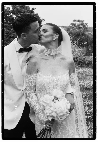 Wedding kiss portrait in a black and white wedding photo, bride in lace dress and veil holding rose bouquet as groom in tux kisses her outdoors