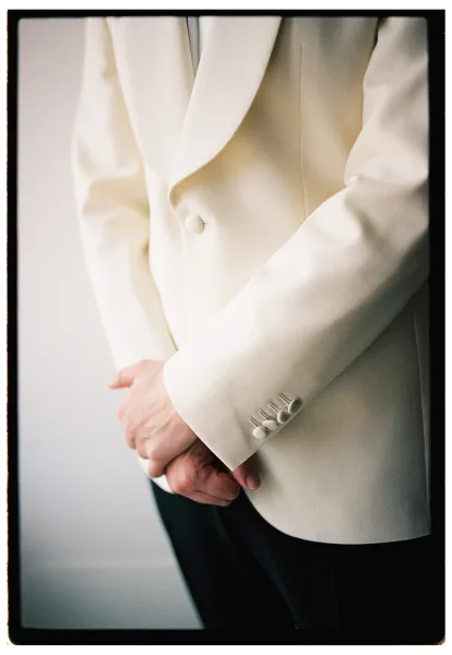 Groom portrait in a white tuxedo jacket with satin shawl lapel and cuff buttons, hands clasped by a neutral wall in soft window light