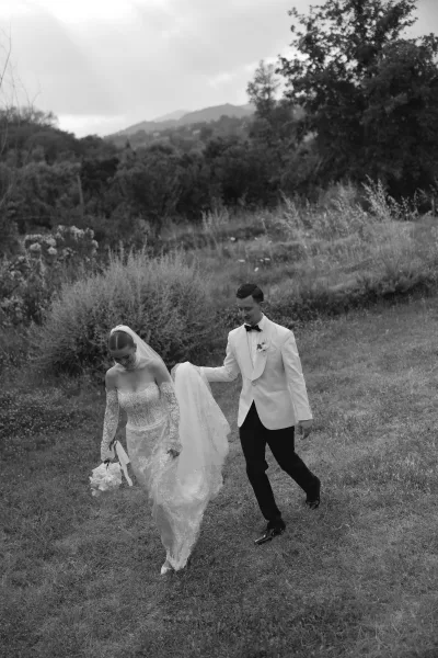 Couple portrait in a black and white wedding photo, bride in lace long sleeves with veil and bouquet, groom lifting her train in a meadow