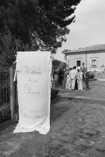 Wedding welcome sign in calligraphy lettering on a hanging fabric banner with ribbon ties along a stone garden path near the villa entrance