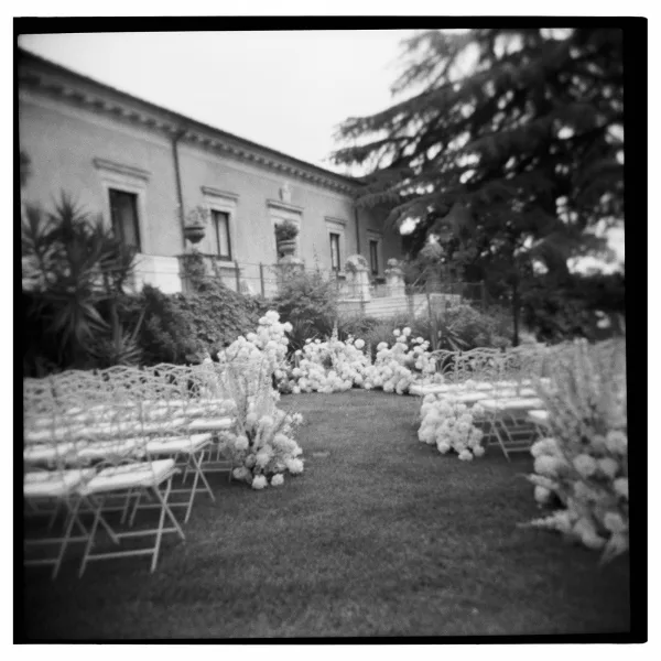 Outdoor ceremony setup with garden wedding ceremony aisle lined by white florals, folding chairs, and a runner on a green lawn by a villa