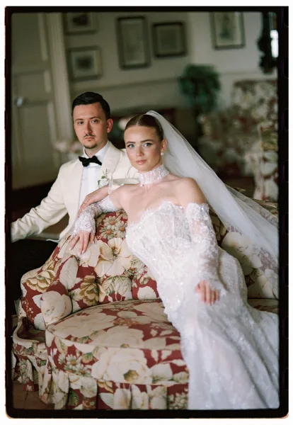 Couple portrait of bride and groom sitting on a floral sofa, bride in veil and lace gloves leaning on groom in white tux indoors