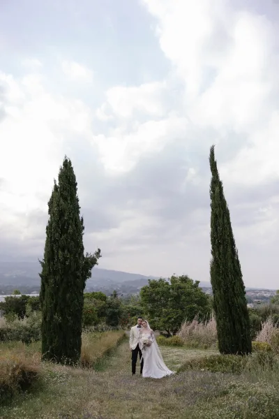 Couple portrait of bride and groom standing full length outdoors, bride in off-shoulder dress with veil and bouquet, hills and cypress trees behind